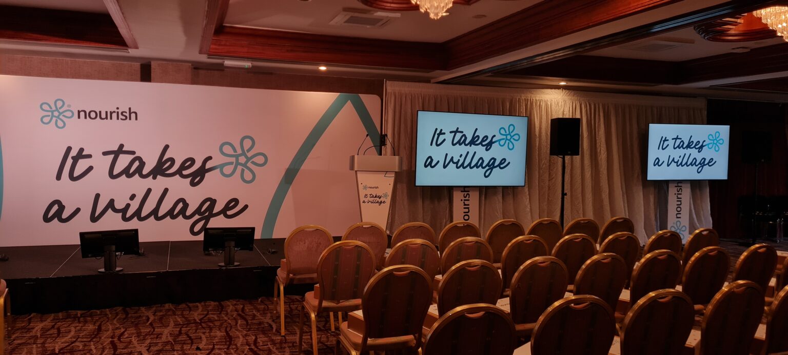 A photo of a stage with "It takes a village" on signage and monitors to the side. There are chairs in a conference layout facing the stage.